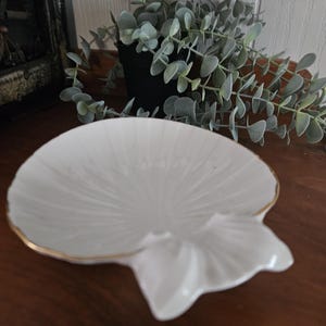May include: A white ceramic dish shaped like a seashell with a delicate gold-painted rim. The surface of the dish features embossed radial ridges that mimic the natural texture of a scallop shell, creating a clean and elegant appearance.