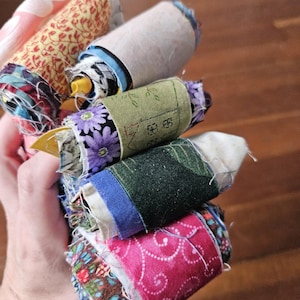 May include: A collection of fabric scraps rolled into small cylinders, featuring a variety of patterns including purple flowers, green foliage, pink swirls, and geometric designs. The rolls are bundled together, displaying frayed edges and diverse colours.