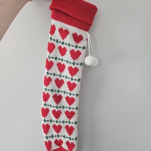 以下が含まれることがあります： 赤い袖口とつま先、赤いハートと緑のアクセント模様の白いボディが特徴の、ニットのクリスマスストッキング。 ストッキングには吊り下げ用のループと2つの白いポンポンが付いています。