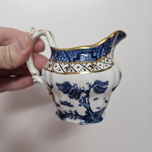 Puede incluir: Un pequeño creamer blanco con un patrón de sauce azul y dorado. El creamer tiene un borde dorado y una banda dorada alrededor del medio.