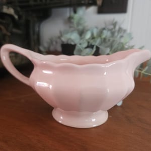 Peut inclure: Une saucière en céramique rose pâle dotée d'un bord festonné, d'une anse incurvée et d'une base sur pied. Le récipient présente une finition lisse et brillante avec des détails cannelés verticaux le long du corps, formant un objet de service.