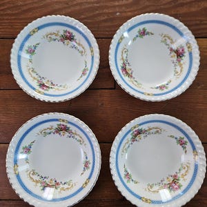Peut inclure: Quatre assiettes en céramique blanche avec une bordure bleue et un motif floral. Les assiettes ont un bord festonné et une bordure dorée. Le motif floral comprend des fleurs roses, violettes et jaunes.