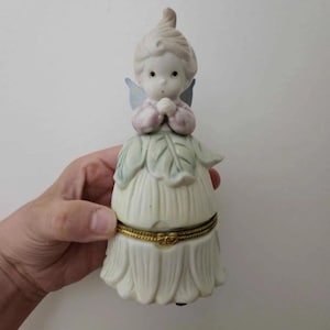 Puede incluir: Una figurita de porcelana de un hada con alas, que lleva un vestido rosa y una falda de hojas verdes. El hada tiene las manos juntas en oración. La figurita es una caja de joyas con una tapa con bisagras de oro.