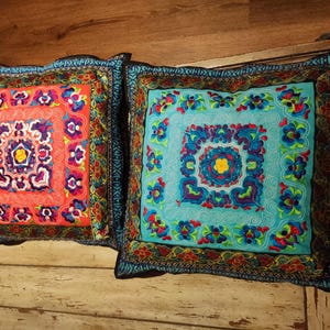 Peut inclure: Deux coussins brodés avec des motifs floraux complexes. Le coussin de gauche a un fond orange vif avec des accents bleus, roses et jaunes. Le coussin de droite a un fond turquoise avec des accents bleus, roses et jaunes.