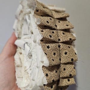 Può includere: Un ornamento in tessuto fatto a mano con bordi a strati e arruffati. L'ornamento presenta un tessuto beige con motivi a stella neri e strati di tessuto bianco e crema. I bordi sono sfilacciati, dando un aspetto rustico e fatto in casa.