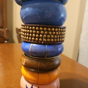 Op de afbeelding: Een stapel kleurrijke armbanden in tinten blauw, bruin, oranje en roze. De armbanden zijn gemaakt van verschillende materialen, waaronder hout en mogelijk hars, en hebben decoratieve elementen zoals studs en geschilderde ontwerpen. De armbanden zijn op elkaar gestapeld.