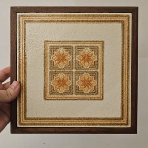 May include: Square decorative tile with a floral design in shades of orange, brown, and cream, set within a dark brown wooden frame. The tile features four floral patterns in the centre, surrounded by a patterned border.