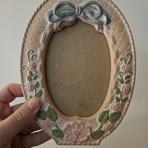 May include: Oval photo frame in beige, decorated with a light blue bow and floral details. Pink roses and green leaves embellish the frame, giving it a vintage look. The frame is held by a hand.