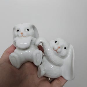 May include: Two white ceramic bunny ornaments. One bunny is sitting with its arms up, and the other is reclining with one ear up. Both bunnies have black eyes and pink noses. The ornaments are held in a hand.