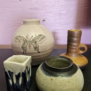 May include: A collection of four ceramic vessels including a round beige vase with a butterfly imprint, a tall brown textured candle holder with a side loop, a small square white and blue drip-glaze container, and a speckled beige bowl.