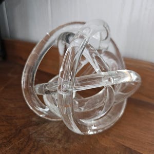 Puede incluir: Escultura decorativa de nudo de vidrio transparente. La escultura está hecha de tubos de vidrio curvados que se cruzan y entrelazan para formar una forma de nudo. El vidrio es transparente, permitiendo el paso de la luz, y se encuentra sobre una superficie de madera.