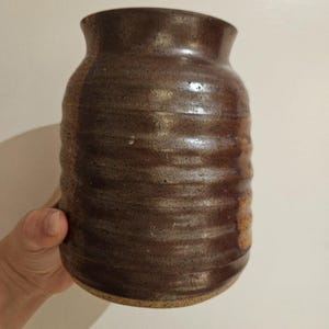 May include: A handcrafted, brown ceramic vase with a textured, ridged exterior. The vase has a wide, rounded body that tapers slightly towards the top, with a narrow opening. The earthy tones and unique shape suggest a rustic, artisanal aesthetic.
