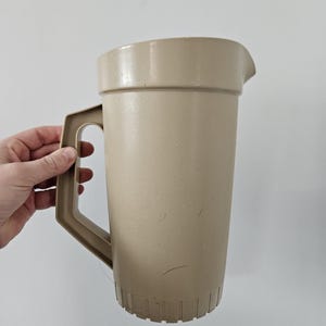 Può includere: Una brocca di plastica beige con un'ampia maniglia e un beccuccio. La brocca ha un corpo cilindrico e una base scanalata. La maniglia è rettangolare e attaccata al lato della brocca. La brocca è tenuta da una mano.