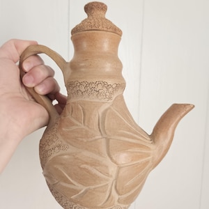 May include: A light brown ceramic teapot with a textured lid and a handle. The body features carved leaf patterns and intricate dotted detailing around the base and neck. The surface has a matte finish with a natural clay appearance.