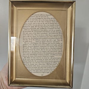 May include: A rectangular gold-toned metal frame containing a page of printed text with an oval-shaped cutout. The visible text discusses Thomas Jefferson, his retirement, and the Library of Congress, ending with the page number 357 at the bottom.