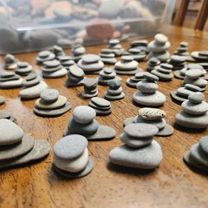 May include: A collection of numerous small, smooth, grey and tan river stones arranged into multiple individual stacks of varying heights. The stones are rounded and flat, creating small towers scattered across a wooden surface.