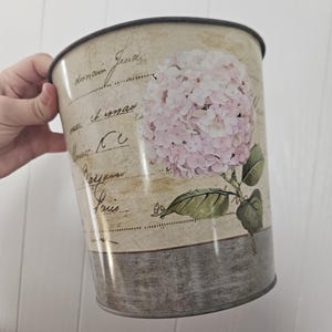 May include: A cylindrical metal waste bin featuring a cream-coloured background with vintage-style cursive French script. The design includes a large, detailed illustration of a pale pink hydrangea flower with green leaves and a dark grey base.