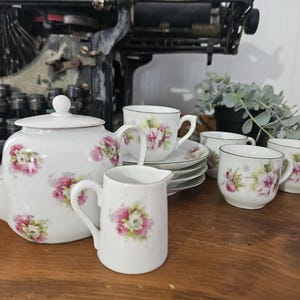 以下が含まれることがあります： ピンクの花柄が施された白い磁器のティーセット。 ティーポット、クリーマー、カップ、ソーサーが含まれています。 ティーセットは木製の表面に置かれ、背景にはヴィンテージのタイプライターがあります。