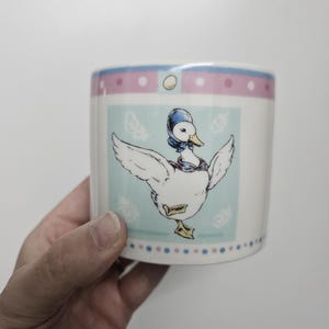 以下が含まれることがあります： 漫画のアヒルのデザインが施された白いセラミックマグ。アヒルは青い帽子とスカーフを身につけ、翼を広げています。マグカップには、水色とピンクのドット模様のボーダーがあります。アヒルは水色の正方形の上にいます。