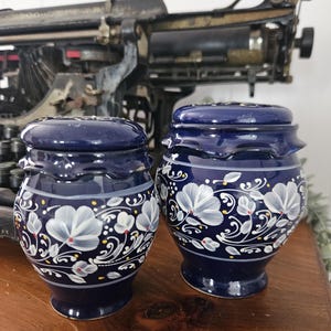Puede incluir: Dos tarros de cerámica azul cobalto a juego con tapas. Cada tarro está decorado con diseños florales blancos y detalles en amarillo y gris. Los tarros están sobre una superficie de madera, con una máquina de escribir vintage al fondo.