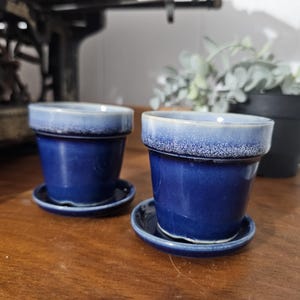 May include: Two matching blue ceramic plant pots with saucers. The pots have a gradient effect, transitioning from a lighter blue rim to a darker blue body. They sit on a wooden surface, with a black pot and greenery in the background.