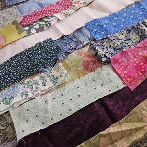 Pode incluir: Variedade de tiras de tecido em várias cores e padrões. Inclui desenhos florais, de bolinhas e geométricos em tons de azul, rosa, verde e roxo. Ideal para projetos de colchas ou artesanato.