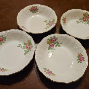 Puede incluir: Cuatro cuencos de porcelana blanca con bordes festoneados y ribete dorado. Cada cuenco presenta un diseño floral con rosas rosas y amarillas y hojas verdes. Los cuencos están dispuestos sobre una superficie marrón oscuro.