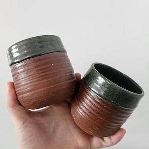 May include: Two handmade ceramic cups. The cups are brown with a ribbed texture on the lower portion and a dark green glaze on the rim. The cups are held in a hand against a white background.