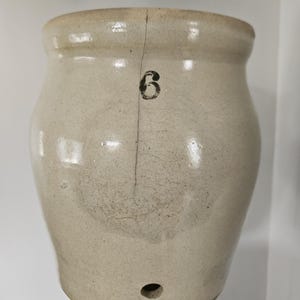 May include: A beige stoneware crock featuring a prominent vertical crack running down the centre. The vessel displays a black printed number 6 near the top rim and a small circular hole located near the base, showcasing a smooth, glazed finish.