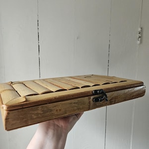 May include: A rectangular bamboo box with a hinged lid and a black metal clasp. The box is held in a hand, showcasing its natural wood tones and textured surface. The lid is made of parallel bamboo strips.