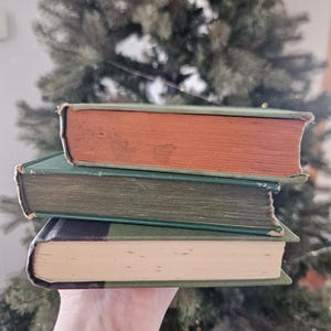 May include: Three vintage books stacked. The top book has a reddish-brown cover, the middle book is green, and the bottom book is green and black. The pages are aged and show signs of wear and tear.
