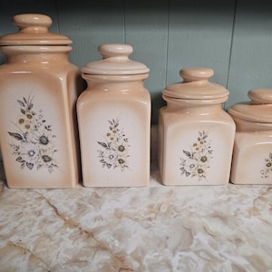 May include: A set of four square ceramic canisters in graduated sizes, featuring a light peach glaze and a central floral decal of white and yellow flowers with grey leaves. Each canister has a matching lid with a rounded knob handle.