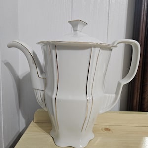 May include: White porcelain teapot with a lid, featuring gold trim and vertical gold stripes. The teapot has a curved spout and a handle. The design is classic and elegant, suitable for serving tea.