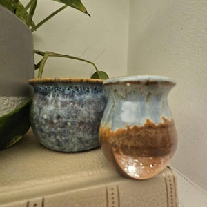 May include: Two small ceramic pots with a blue and brown speckled glaze. The pots are on a book, with a plant behind them. The pots have a unique shape and texture, suitable for small plants or decorative purposes. The pots are handmade.