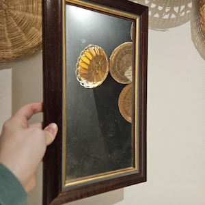 以下が含まれることがあります： 壁に掛けられた、濃い木製のフレームと金色の縁取りのある鏡。鏡には、籐のバスケットと金色の金属製のボウルが映っています。