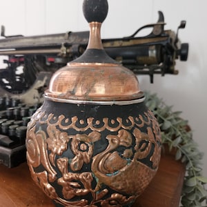 May include: A decorative copper and black jar with a lid. The jar features embossed floral and bird designs. The lid has a copper base and a black finial. The jar is displayed on a wooden surface.