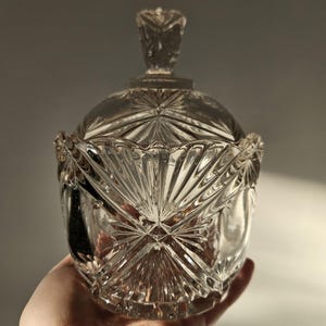 Könnte beinhalten: Eine transparente Bonbonniere aus geschliffenem Glas mit Deckel. Die Schale hat ein Sternmuster auf dem Boden und dem Deckel, mit einem dekorativen Knauf. Das Glas hat ein facettiertes Design.
