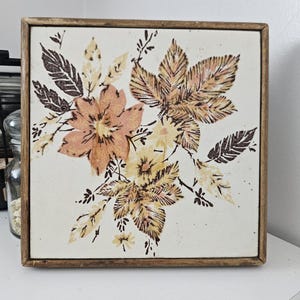 May include: A framed tile with a floral design featuring brown, orange, and yellow flowers and leaves on a white background.