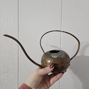 May include: A small, round, hammered copper watering can with a long, slender, curved spout and a thin, arched handle. The top surface of the metal body features distinct patches of blue and green oxidation, showing a weathered patina.