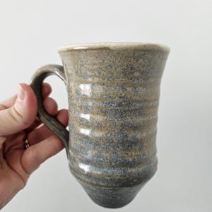 May include: A handcrafted ceramic mug with a textured, ridged pattern. The mug has a neutral colour scheme with shades of beige, grey, and a darker base. The handle is integrated. This stoneware mug is suitable for tea or coffee.