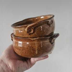 May include: Two handmade, brown ceramic bowls stacked. Each bowl has a rounded shape, dark brown rim and two small handles. The bowls have a speckled texture and a glossy finish. Ideal for serving soups or stews.