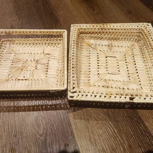May include: Two woven wicker trays, one slightly larger than the other, both with a square shape and a natural beige colour.