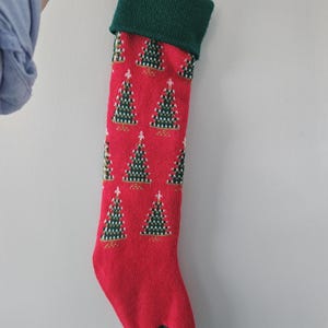 Peut inclure: Chaussette de Noël rouge avec un revers et un talon verts. La chaussette est ornée de motifs répétés de sapins de Noël verts et blancs. Une boucle verte est attachée pour l'accrocher.
