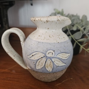 May include: A small, handmade ceramic jug with a handle. The jug features a light blue band with a white floral design. The top and base are speckled with brown, and the handle is curved.