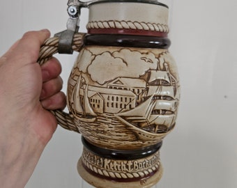 May include: A ceramic beer stein with a pewter lid and a rope-like design around the top. The stein features a detailed image of a ship sailing into a harbor with buildings in the background. The text "Headed Kettch Barkentine" is visible on the bottom of the stein.