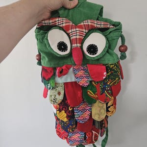 Peut inclure: Un sac à dos fait à la main en forme de hibou. Il présente un corps vert, des accents à carreaux rouges et blancs et de grands yeux blancs avec des pupilles noires. Le corps du hibou est orné d'écailles en tissu colorées et à motifs, et il a une sangle verte.
