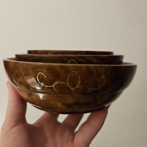 May include: A set of three wooden bowls in different sizes. The bowls are a deep brown colour with a glossy finish. Each bowl has gold-coloured decorative details. The bowls are held in a hand, showcasing their varying sizes and the natural wood grain.