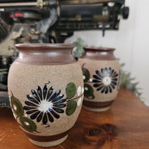May include: Two small, round ceramic vases. The vases have a tan base and brown rim, each decorated with a blue and white floral design and green leaves. They sit on a wooden surface, with a vintage typewriter in the background.