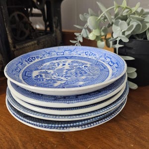 Puede incluir: Una pila de cinco platos de cerámica azules y blancos. Cada plato presenta un diseño detallado de paisaje en azul sobre un fondo blanco. Los platos están apilados sobre una superficie de madera, mostrando una estética clásica y vintage.