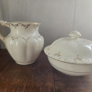 May include: Cream-coloured ceramic jug and lidded dish with gold trim. The jug has a handle and a flared rim. The lidded dish has a rounded shape with a lid and a decorative gold pattern. Both pieces are on a wooden surface.